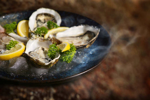 Assiette bleue avec des huîtres fraîches ouvertes garnies de persil et quartiers de citron sur lit de glace.